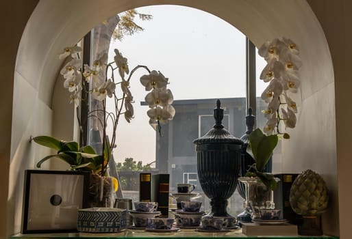 Beautiful antique tea cups and Ceramic jar collection with White orchid flower pot displayed in the window. Selective focus.