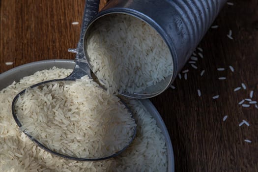 Thai jasmine rice pouring from old steel can with steel dipper on bowl on an old rustic wooden background. Healthy food and diet concept. Selective focus.