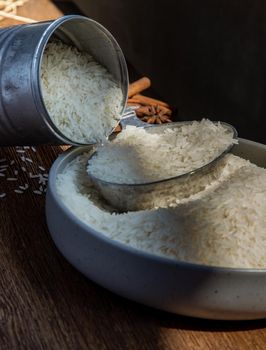 Thai jasmine rice pouring from old steel can with steel dipper on bowl on an old rustic wooden background. Healthy food and diet concept. Selective focus.