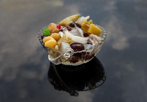 Traditional chinese iced sweet dessert is delicious here are many grains including Sweet potato balls, Grass jelly, Red beans, Ginkgo, Taro balls, lotus seeds, millet, persimmon, longan and boiled flour Topping with boil sugar syrup in Glass bowl. Selective focus.