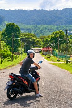 A tourist guy on a motorbike stands in the middle of the road overlooking the jungle. Tropical exotic island in Asia.