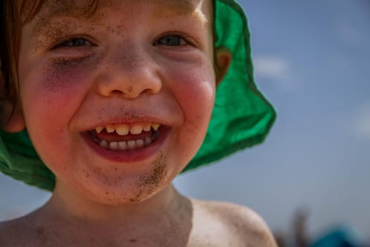 Portrait of a cute, redhead, blue-eyed baby boy in the beach wearing a green hat and with his face covered in sand