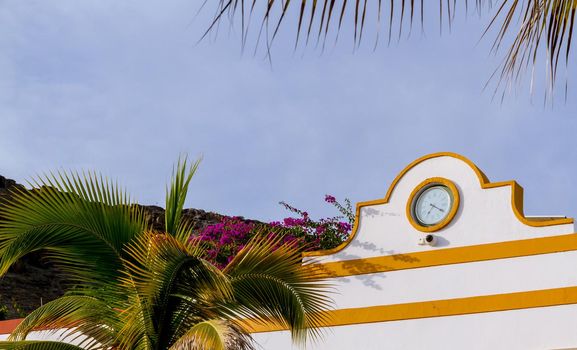 Detail of a clock in a white wall with yellow details by a palm tree on a sunny day