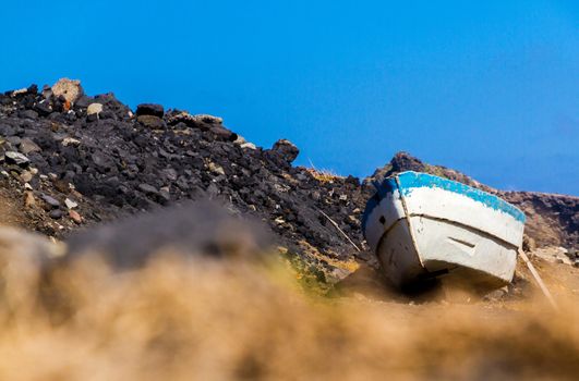 Low-angle view of a small white and blue skiffle on dry land by some black rocks on a sunny day