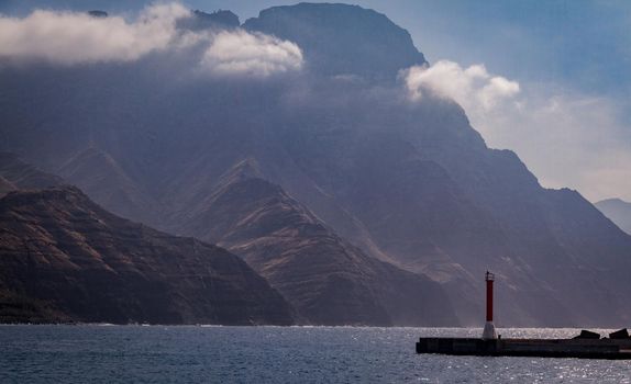 Detail of a small red and white beacon by the sea with big mountains covered by clouds in teh background