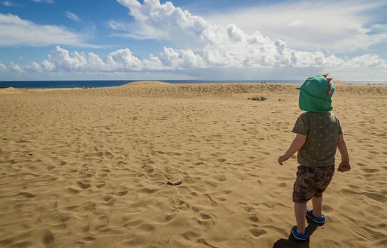 A baby boy with a green hat, t-shirt and shorts walking in an empty sandy beach with teh sea at background