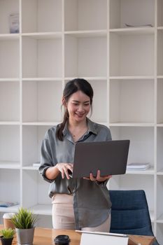 Young asian woman standing and holding laptop browsing internet at home office
