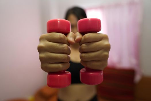 Asian woman training to lift pink dumbbells for building muscle, concept of staying healthy by weight loss and recreation at their own residence.