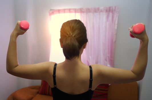 Asian women training to lift pink dumbbells for building muscle, the concept of staying healthy by weight loss and recreation at their residence.