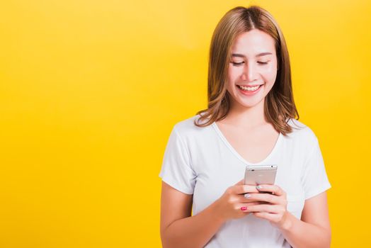 Asian Thai portrait happy beautiful cute young woman smile standing playing game or writing SMS message on smartphone looking to the phone, studio shot isolated on yellow background with copy space