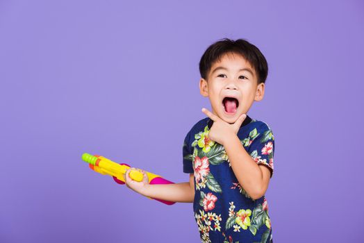 Thai kid funny hold toy water pistol and smiling, Happy Asian little boy holding plastic water gun, studio shot isolated on purple background, Thailand Songkran festival day national culture concept