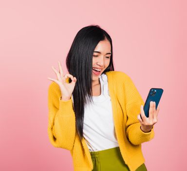 Happy Asian portrait beautiful cute young woman excited holding mobile phone and gesturing ok sign, studio shot isolated on pink background, Thai female making finger symbol on smartphone