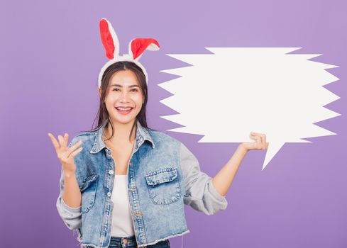 
Happy Easter Day. Beautiful young woman smiling excited wearing rabbit ears and denims holding empty speech bubble, Portrait female looking at camera, studio shot isolated on purple background