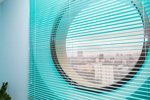 Office interior with a round window closed by blue blinds. View of the city from the window