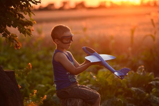 A boy with pilot's glasses is playing with an airplane. The child imagines himself as an aviator pilot. Outdoor games in summer at sunset.