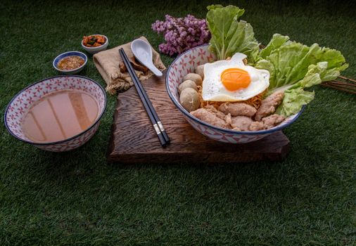 Boil egg noodles with pork ball, minced pork, fried egg, vegetables and soups in bowl and chopstick with spoon and timber background. Thai noodle soup.