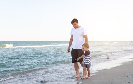 Young Caucasian dad with little son walk warm summer day along the sea coast. Summer family vacation concept. Friendship father and son. Happy childhood on the beach with parent. free space for text