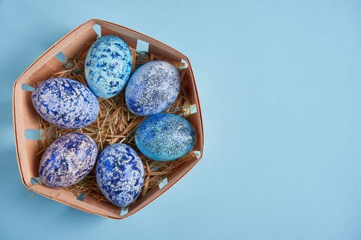 Blue chicken eggs lie in round wooden basket which stands on a blue paper background. Easter background. Seasonal holiday flat lay with free space for text. Classic blue color of the year 2020