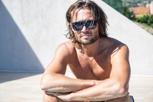 The handsome brutal man in sunglasses with a long hair and naked torso sits near the pool, a sports suntanned body, sunglasses with a blue frame, he is red blue swimming shorts, sunny day