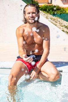 The handsome brutal man with a long hair and naked torso sits near the pool, splashes with water, he has fun, a sports suntanned body, he is red blue swimming shorts