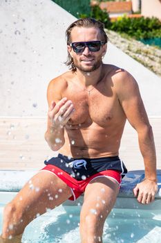 The handsome brutal man in sunglasses with a long hair and naked torso sits near the pool, a sports suntanned body, sunglasses with a blue frame, he is red blue swimming shorts, sunny day