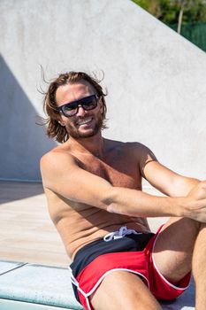 The handsome brutal man in sunglasses with a long hair and naked torso sits near the pool, is looking a camera, big smile, a sports suntanned body, sunglasses with a blue frame, sunny