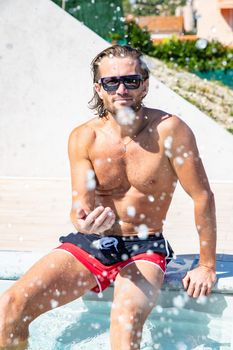 The handsome brutal man in sunglasses with a long hair and naked torso sits near the pool, a sports suntanned body, sunglasses with a blue frame, he is red blue swimming shorts, sunny day