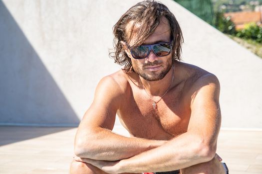 The handsome brutal man in sunglasses with a long hair and naked torso sits near the pool, a sports suntanned body, sunglasses with a blue frame, he is red blue swimming shorts, sunny day