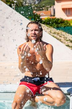 The handsome brutal man with a long hair and naked torso sits near the pool, splashes with water, he has fun, a sports suntanned body, he is red blue swimming shorts