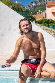 The handsome brutal man with a long hair and naked torso sits near the pool, splashes with water, he has fun, a sports suntanned body, he is red blue swimming shorts