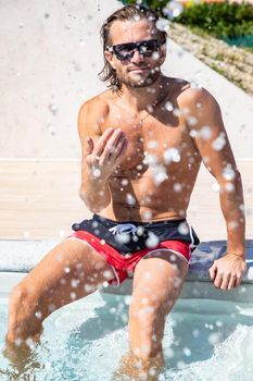 The handsome brutal man with a long hair and naked torso sits near the pool, splashes with water, he has fun, a sports suntanned body, he is red blue swimming shorts