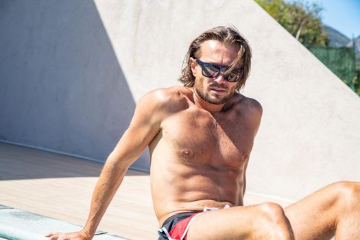 The handsome brutal man in sunglasses with a long hair and naked torso sits near the pool, a sports suntanned body, sunglasses with a blue frame, he is red blue swimming shorts, sunny day