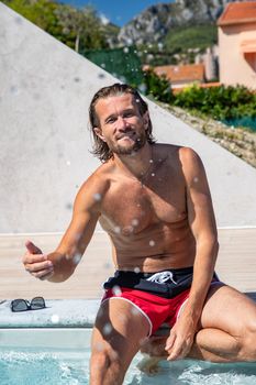 The handsome brutal man with a long hair and naked torso sits near the pool, splashes with water, he has fun, a sports suntanned body, he is red blue swimming shorts