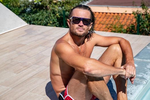 The handsome brutal man in sunglasses with a long hair and naked torso sits near the pool, a sports suntanned body, sunglasses with a blue frame, he is red blue swimming shorts, sunny day