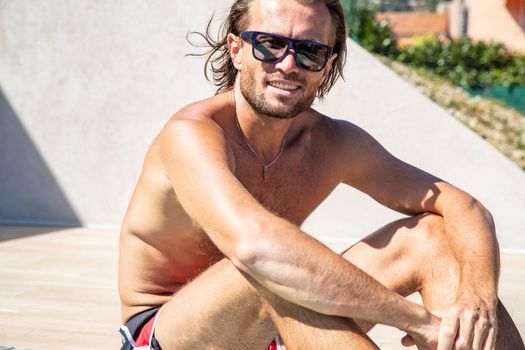 The handsome brutal man in sunglasses with a long hair and naked torso sits near the pool, a sports suntanned body, sunglasses with a blue frame, he is red blue swimming shorts, sunny day