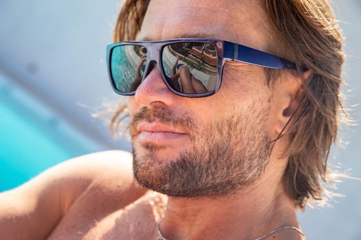 The handsome brutal man in sunglasses with a long hair and naked torso sits near the pool, a sports suntanned body, sunglasses with a blue frame, he is red blue swimming shorts, sunny day