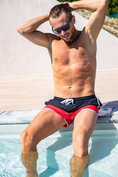 The handsome brutal man in sunglasses with a long hair and naked torso sits near the pool, a sports suntanned body, sunglasses with a blue frame, he is red blue swimming shorts, sunny day