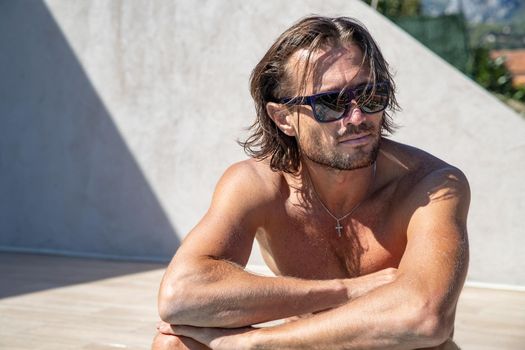 The handsome brutal man in sunglasses with a long hair and naked torso sits near the pool, a sports suntanned body, sunglasses with a blue frame, he is red blue swimming shorts, sunny day