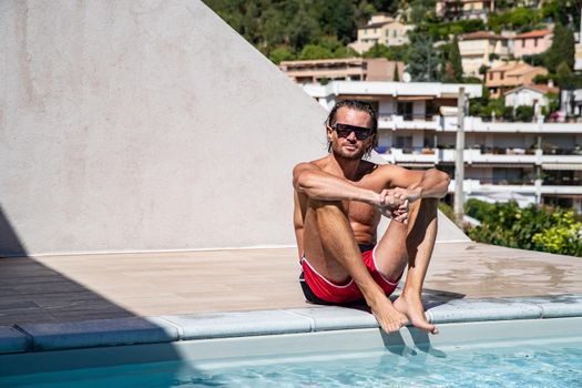 The handsome brutal man in sunglasses with a long hair and naked torso sits near the pool, a sports suntanned body, sunglasses with a blue frame, he is red blue swimming shorts, sunny day