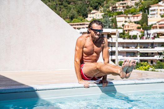 The handsome brutal man in sunglasses with a long hair and naked torso sits near the pool, a sports suntanned body, sunglasses with a blue frame, he is red blue swimming shorts, sunny day