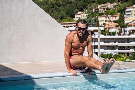 The handsome brutal man in sunglasses with a long hair and naked torso plays sports near the pool, a suntanned fitness body, sunglasses with a blue frame, he is red blue swimming shorts
