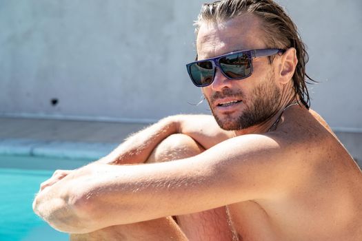 The handsome brutal man in sunglasses with a long hair and naked torso sits near the pool, a sports suntanned body, sunglasses with a blue frame, he is red blue swimming shorts, sunny day