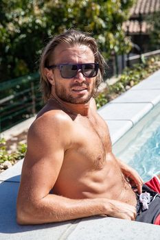 The handsome brutal man in sunglasses with a long hair and naked torso sits near the pool, a sports suntanned body, sunglasses with a blue frame, he is red blue swimming shorts, sunny day