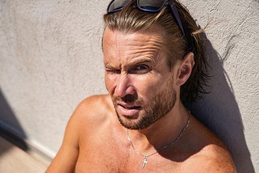 The handsome brutal man in sunglasses with a long hair and naked torso sits near the pool, a sports suntanned body, sunglasses with a blue frame, he is red blue swimming shorts, sunny day