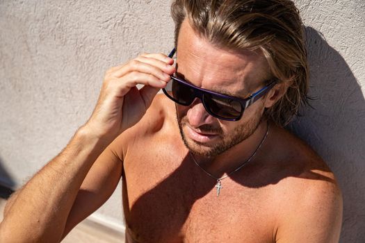 The handsome brutal man in sunglasses with a long hair and naked torso sits near the pool, a sports suntanned body, sunglasses with a blue frame, he is red blue swimming shorts, sunny day