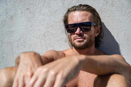 The handsome brutal man in sunglasses with a long hair and naked torso sits near the pool, a sports suntanned body, sunglasses with a blue frame, he is red blue swimming shorts, sunny day