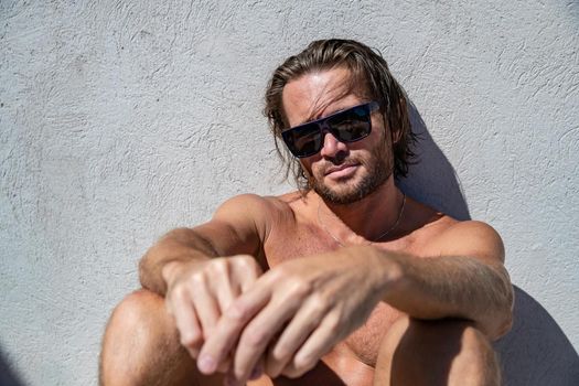 The handsome brutal man in sunglasses with a long hair and naked torso sits near the pool, a sports suntanned body, sunglasses with a blue frame, he is red blue swimming shorts, sunny day