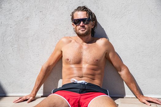 The handsome brutal man in sunglasses with a long hair and naked torso sits near the pool, a sports suntanned body, sunglasses with a blue frame, he is red blue swimming shorts, sunny day