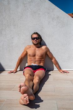 The handsome brutal man in sunglasses with a long hair and naked torso sits near the pool, a sports suntanned body, sunglasses with a blue frame, he is red blue swimming shorts, sunny day