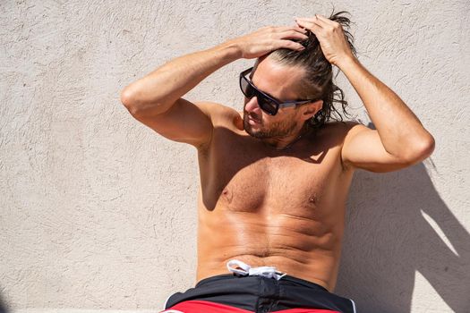 The handsome brutal man in sunglasses with a long hair and naked torso sits near the pool, a sports suntanned body, sunglasses with a blue frame, he is red blue swimming shorts, sunny day